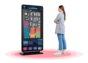 A woman stands with her arms crossed, looking at a large, vertical digital signage screen. The screen displays a colorful floor plan.
